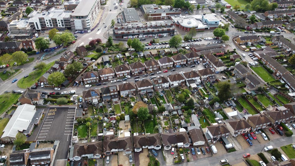 An aerial view of a residential neighbourhood showing a row of semi-detached houses with small, well-maintained gardens at the back, featuring lawns, flowerbeds, and patio areas. The houses have brown